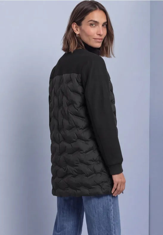 Femme portant une longue veste noire à dos matelassé, regardant par-dessus l'épaule.