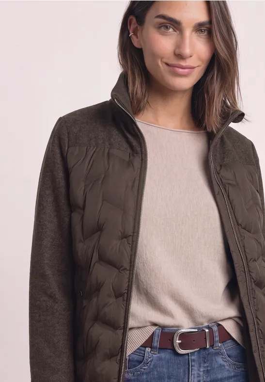 Femme en veste matelassée kaki, pull beige et ceinture marron.