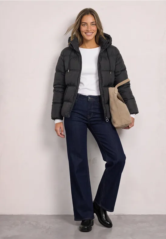 Een glimlachende vrouw draagt een zwarte gewatteerde jas, een wit shirt, wijde jeans en een tas.