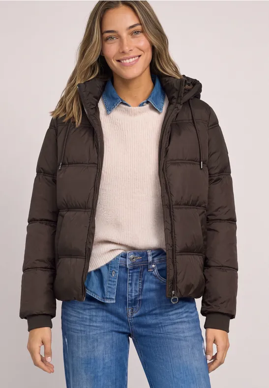 Eine junge Frau lächelt und trägt eine braune Steppjacke über Strickpullover und Jeans.