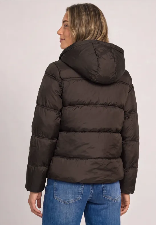 Eine Frau in brauner Steppjacke mit Kapuze und blauen Jeans vor hellem Hintergrund.
