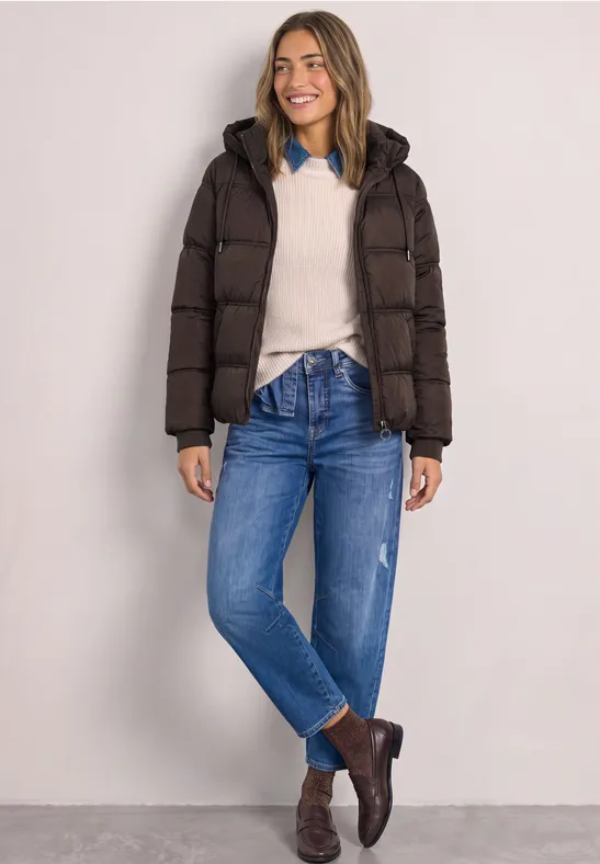 Frau steht lächelnd in dunkler Steppjacke, hellem Pullover, Jeans und braunen Boots vor neutralem Hintergrund.