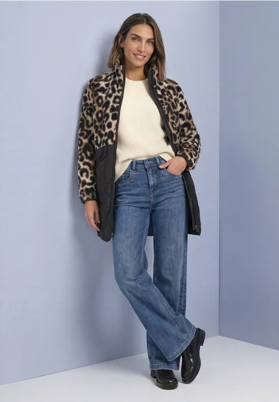 Femme portant un manteau léopard, un pull clair et un jean, posant devant un mur bleu.