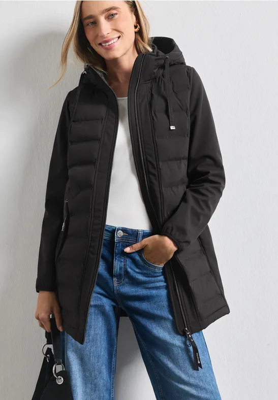 Une femme souriante porte une doudoune noire matelassée à capuche, associée à un pull blanc et un jean.