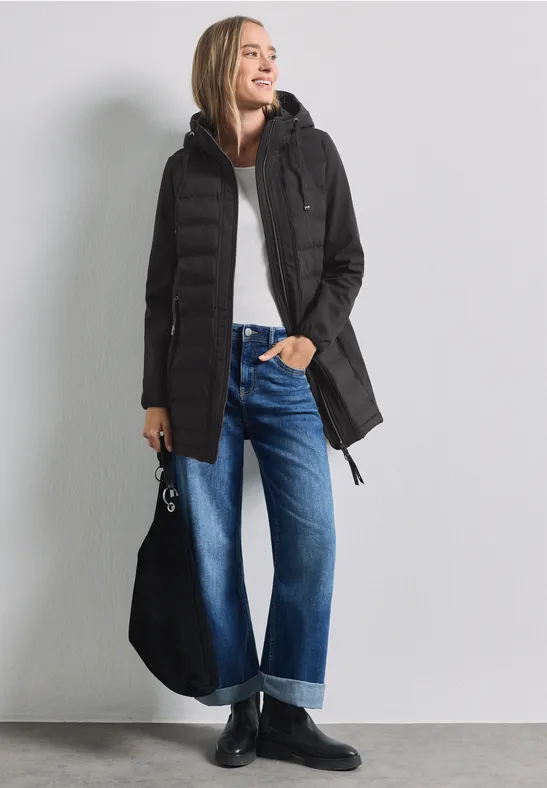 Une femme porte un manteau noir à capuche, un haut blanc, un jean large bleu retroussé et des bottines noires.