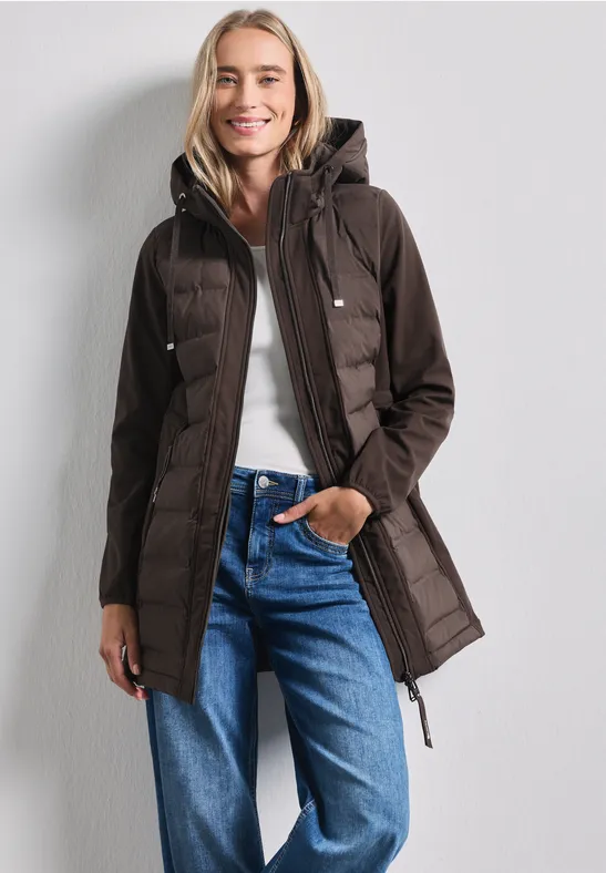 Une femme porte une parka matelassée marron à capuche, un top blanc et un jean bleu.