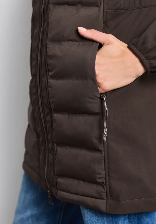 Veste matelassée marron avec main dans la poche zippée, portée sur un jean bleu.