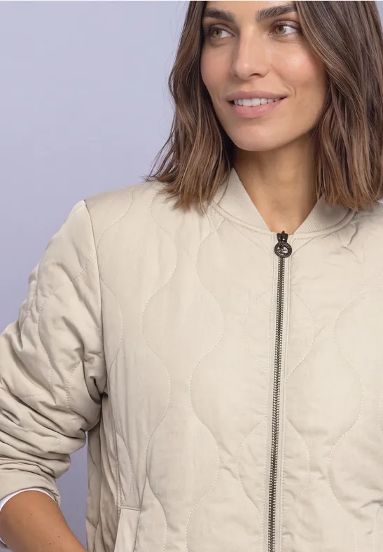 Femme en veste matelassée beige à fermeture éclair et col rond, fond clair.