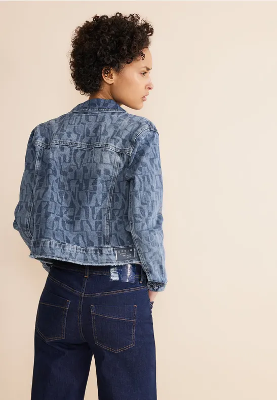 Persoon met krullend haar draagt een spijkerjack met patroon en donkere jeans.