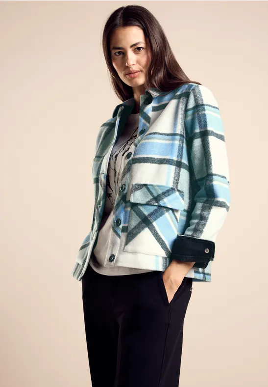 Vrouw met blauw-groen geruit jasje, zwarte broek en licht shirt.
