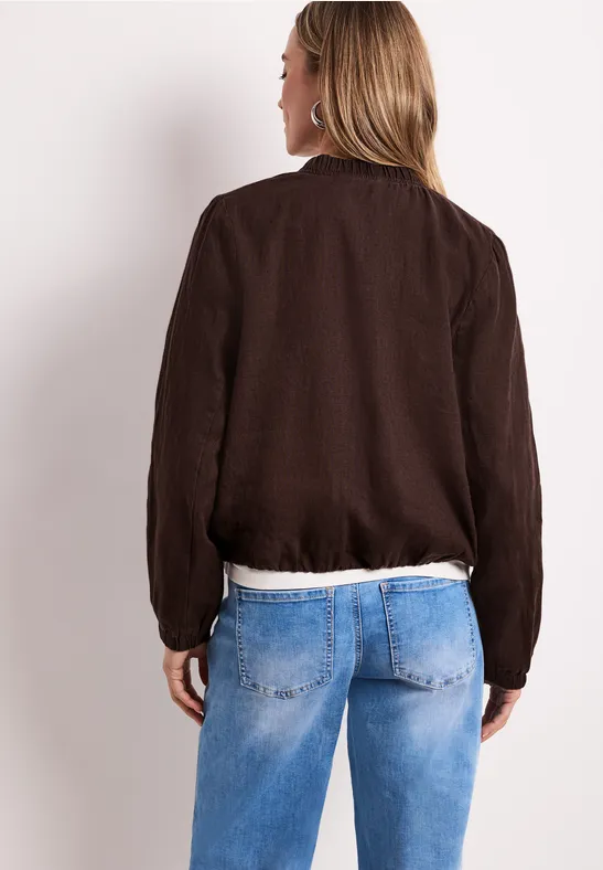 Frau steht Rücken zur Kamera, trägt eine dunkelbraune Jacke über weißem Oberteil und blaue Jeans.