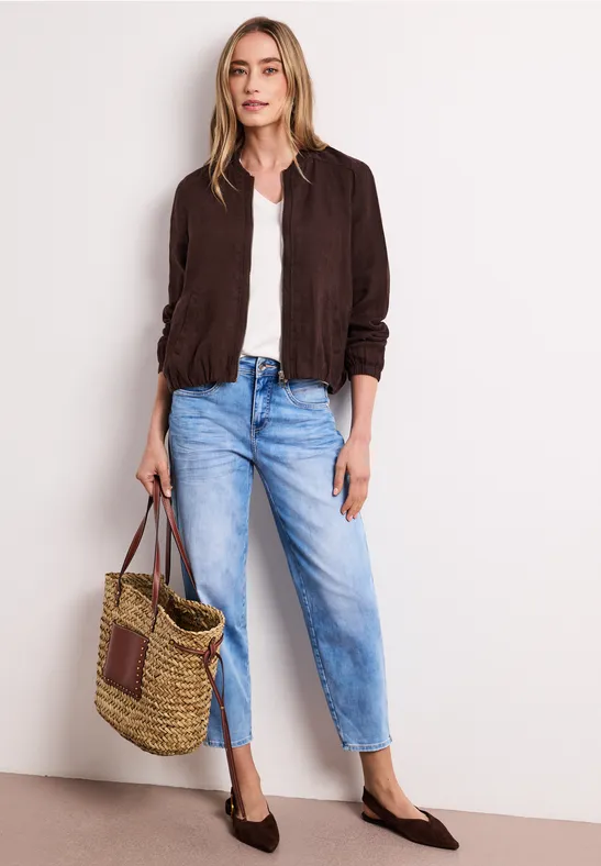 Frau trägt weiße Bluse, dunkelbraune Jacke, helle Jeans, braune Schuhe und Korbtasche.