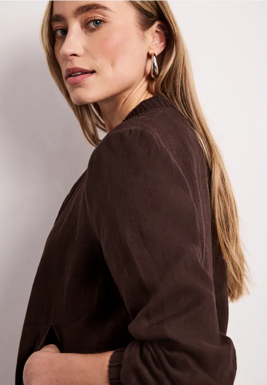 Seitliche Ansicht einer Frau mit braunem Langarmshirt, langen blonden Haaren und Ohrringen vor hellem Hintergrund.