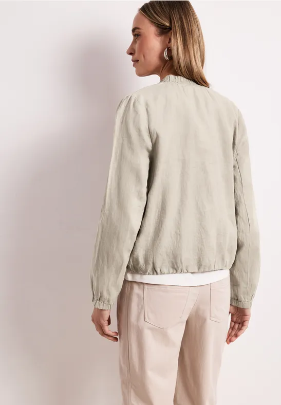 Eine Frau steht Rücken zur Kamera und trägt eine hellbeige Jacke, weiße Bluse und rosafarbene Hose.