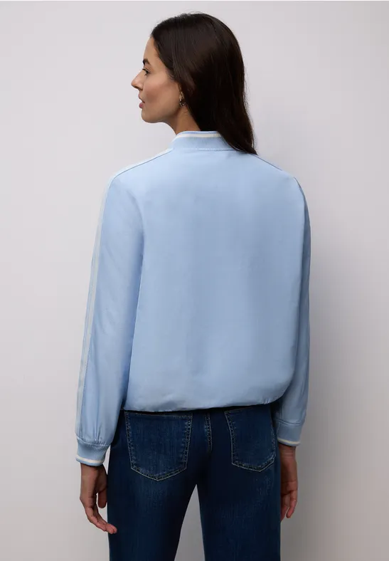 Frau von hinten in hellblauer Bluse und Jeans vor neutralem Hintergrund.
