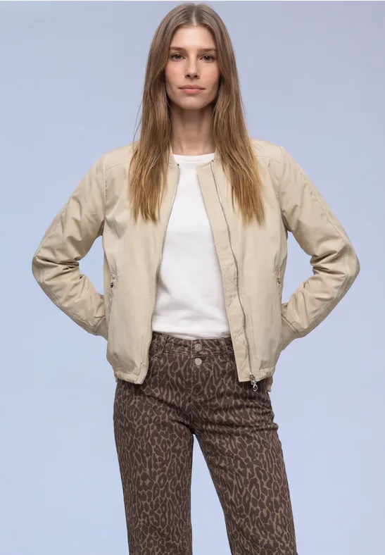 Eine Person trägt eine hellbeige Bomberjacke, weißes T-Shirt und gemusterte braune Hose.
