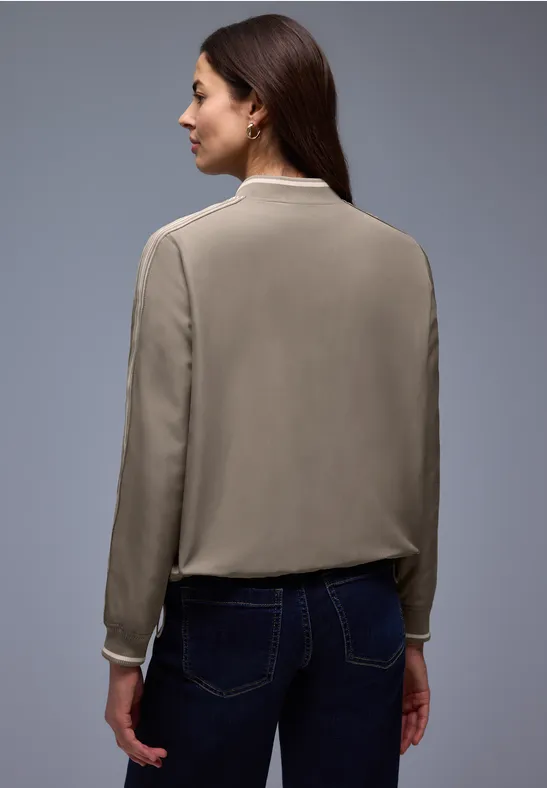 Persoon van achter gezien in een beige blouse met lange mouwen en donkere broek tegen neutrale achtergrond.