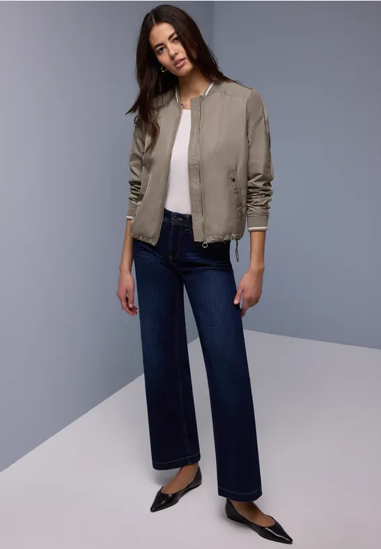 Vrouw in kaki bomberjack, zwart topje, wijde donkere jeans en zwarte puntige ballerina's.