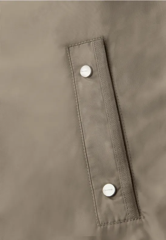 Close-up van een taupekleurige jaszak met twee zilverkleurige drukknoppen.