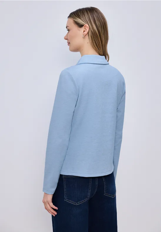 Rückansicht einer Frau in einem hellblauen, langärmeligen Hemd und dunklen Jeans.