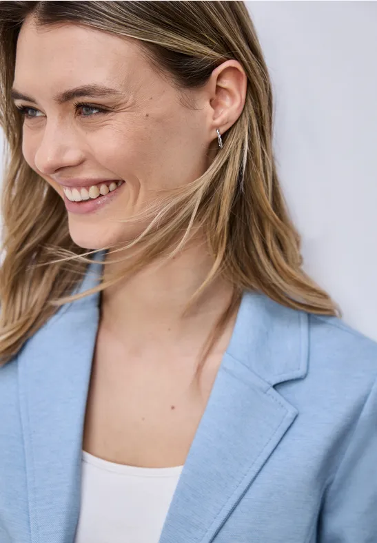 Eine Person mit langen blonden Haaren trägt ein hellblaues Sakko und ein weißes Top.