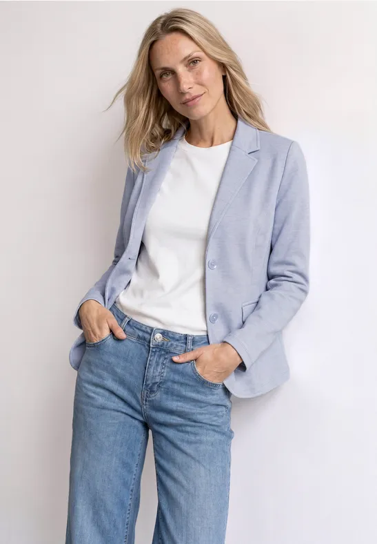 Een persoon draagt een lichtblauw jasje, wit T-shirt en blauwe jeans, staat nonchalant met handen in de zakken.