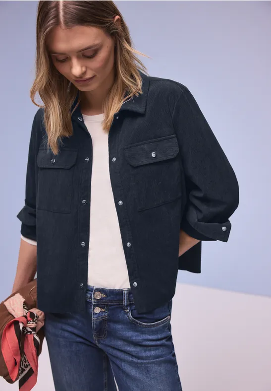 Persoon draagt donker jasje, wit topje en blauwe jeans, houdt rood doek vast tegen lichte achtergrond.