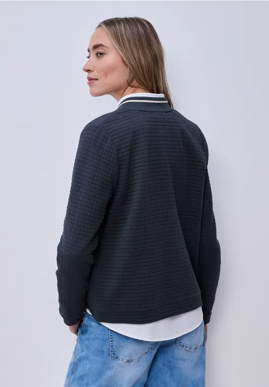 Eine Person mit langen Haaren trägt von hinten gesehen einen dunkelblauen Pullover über einem weißen Hemd und Jeans.