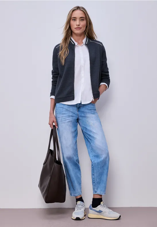 Eine Frau trägt schwarzen Blazer, weiße Bluse, weite Jeans, Sneaker und hält eine große Tasche.