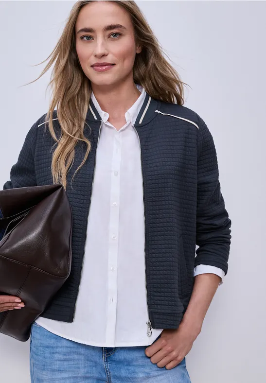 Frau in weißer Bluse, dunkler Strickjacke, blauen Jeans und mit großer brauner Ledertasche.