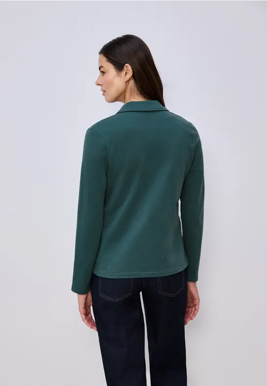 Vrouw van achteren in donkergroene blazer en donkere broek voor lichte achtergrond.