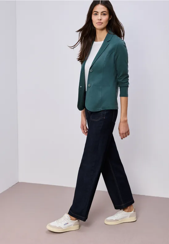 Vrouw in groene blazer, wit shirt, wijde donkerblauwe jeans en witte sneakers.