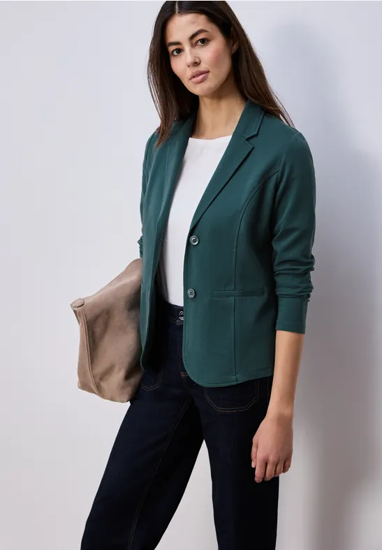 Persoon draagt groene blazer, wit shirt en zwarte broek, houdt een beige tas vast.