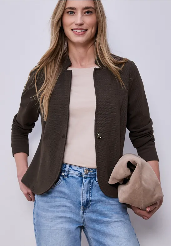 Frau in schwarzer Jacke, beigem Top, Jeans und passender Tasche vor neutralem Hintergrund.