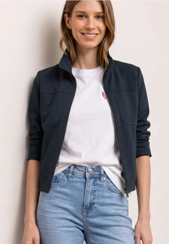 Frau in weißem T-Shirt mit kleinem rotem Logo, dunkelblauem Cardigan und hellblauen High-Waist-Jeans.