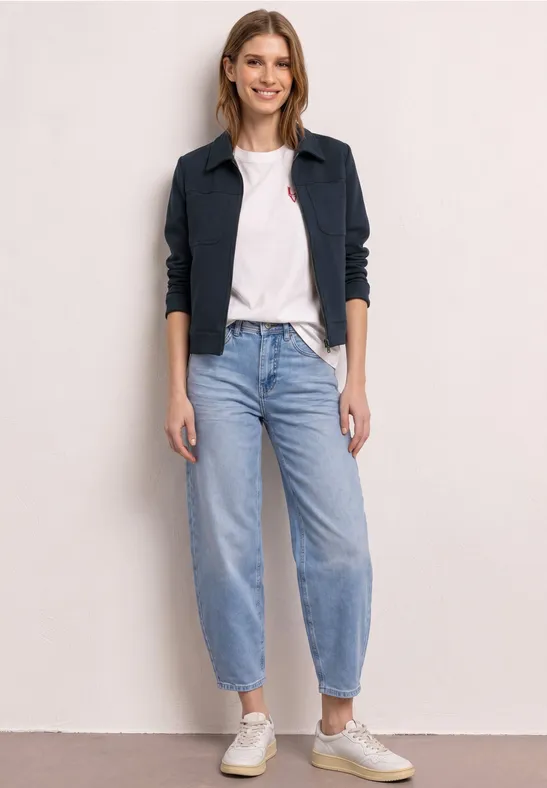Frau in dunkler Jacke, weißem T-Shirt, hellblauen Jeans und weißen Sneakern vor hellem Hintergrund.