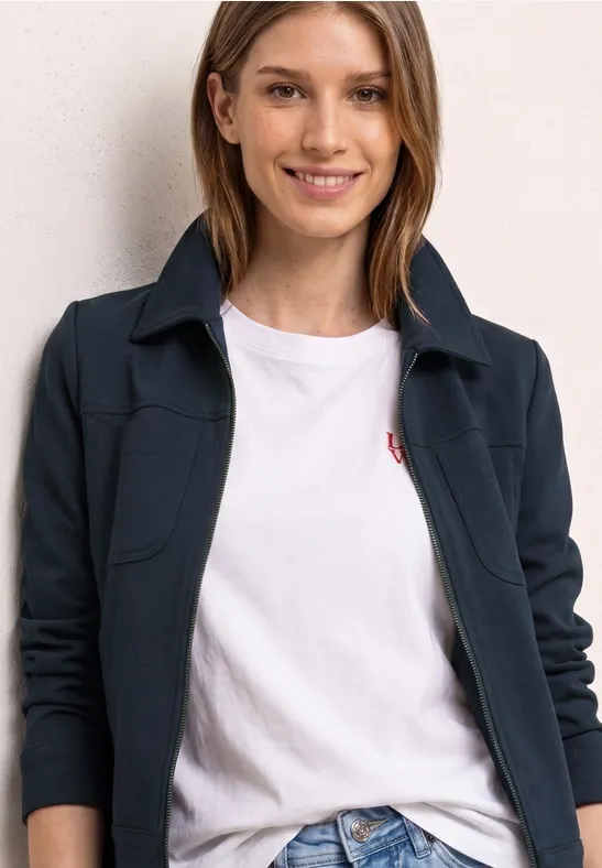 Person trägt eine dunkelblaue Jacke mit Reißverschluss und ein weißes T-Shirt mit kleinem roten Logo.