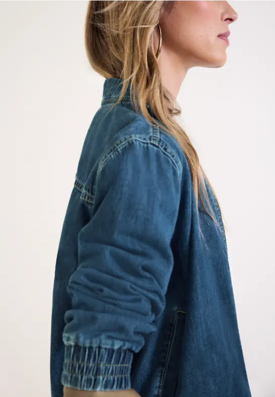 Seitenporträt einer Frau mit langen blonden Haaren und Jeansjacke vor hellem Hintergrund.