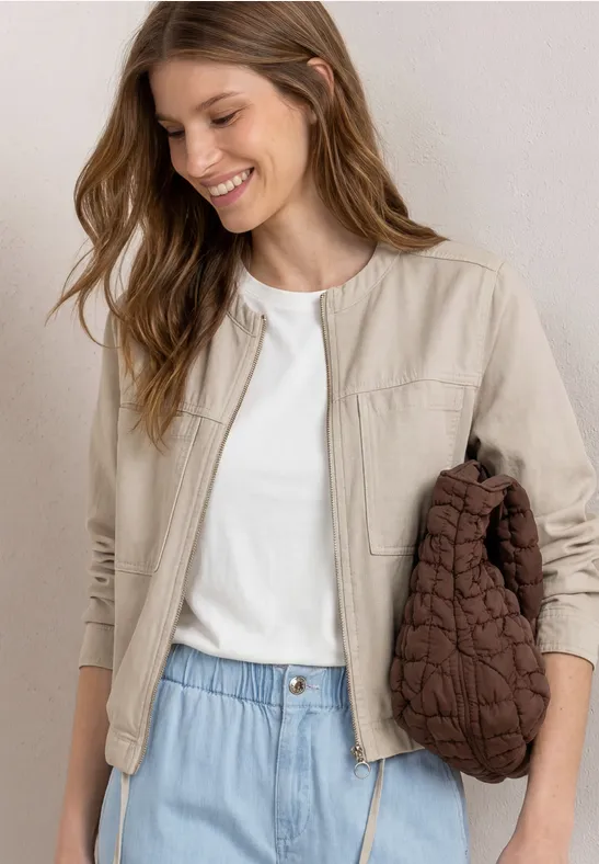 Femme en blouson beige, t-shirt blanc et jean clair, tenant un sac matelassé marron.
