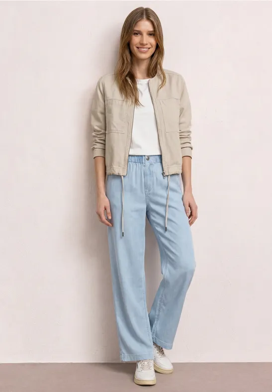 Femme portant une veste en cuir beige, un t-shirt blanc, un jean large bleu et des baskets blanches devant un mur clair.