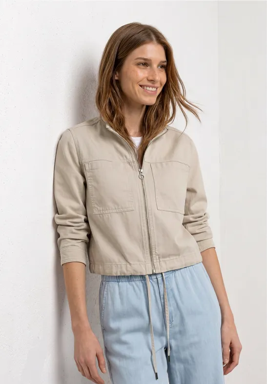 Une femme devant un fond blanc porte une veste couleur sable zippée et un pantalon ample bleu clair.