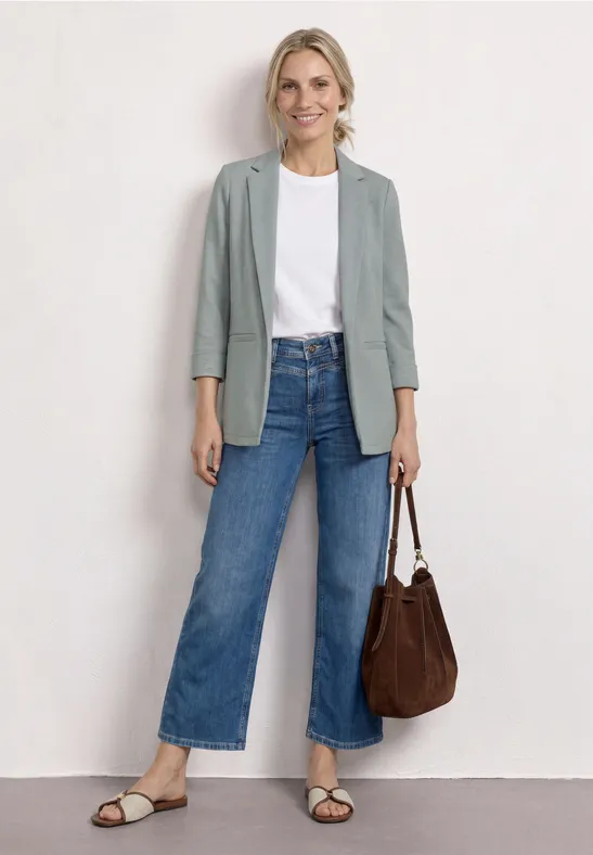 Vrouw in wit T-shirt, grijze blazer, wijde jeans, sandalen en bruine tas.