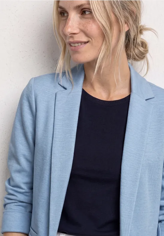 Vrouw met blond knotje draagt lichtblauw colbert en zwart topje voor lichte achtergrond.