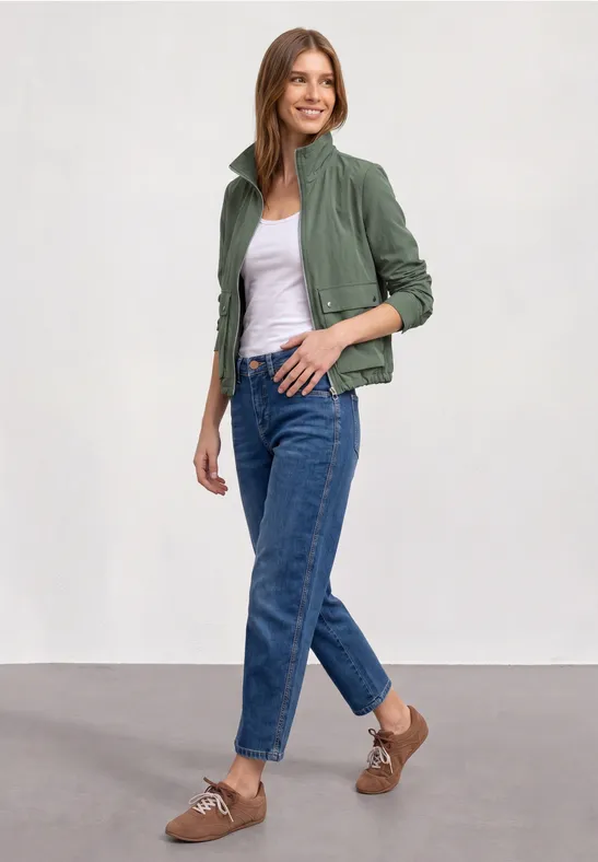 Eine Frau trägt eine grüne Jacke, weißes Top, blaue Jeans und braune Slingback-Schuhe.