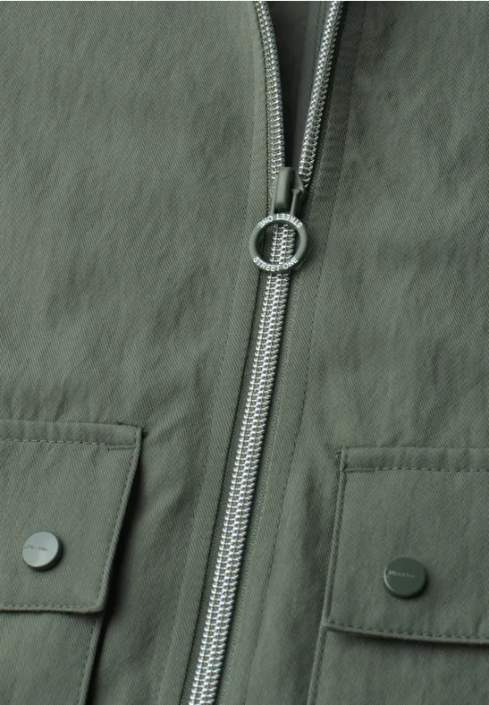 Nahaufnahme einer olivgrünen Jacke mit Reißverschluss und zwei aufgesetzten Taschen mit Metallknöpfen.