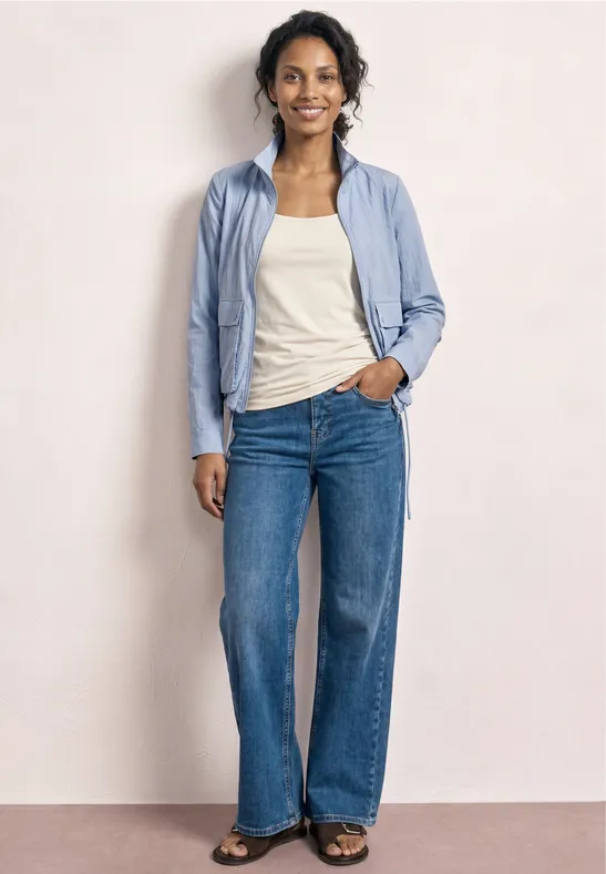 Frau vor hellem Hintergrund, hellblaue Bluse, weißes Top und weite blaue Jeans.