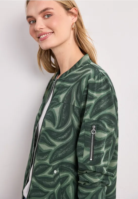 Frau mit blonden Haaren trägt eine grüne Bomberjacke mit psychedelischem Wirbelmuster und lächelt zur Seite.