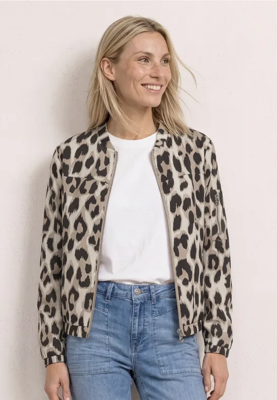 Blonde vrouw draagt een panterprint bomber, wit shirt en blauwe jeans voor een lichte achtergrond.