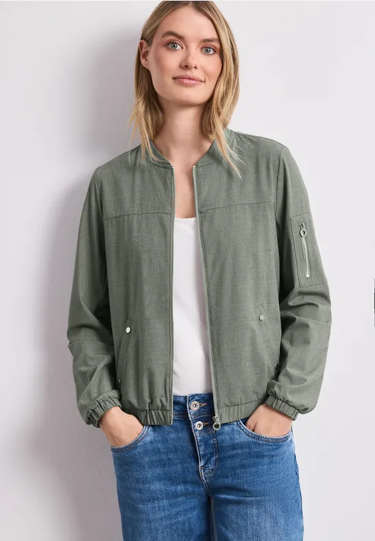 Frau in grüner Bomberjacke, weißem Top und blauen Jeans.