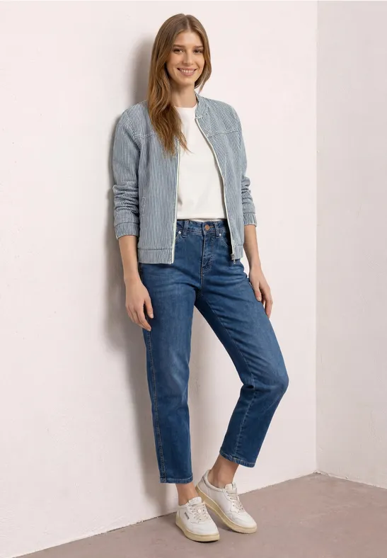 Een vrouw leunt tegen een muur, gekleed in een licht jasje, wit t-shirt, blauwe jeans en witte sneakers.